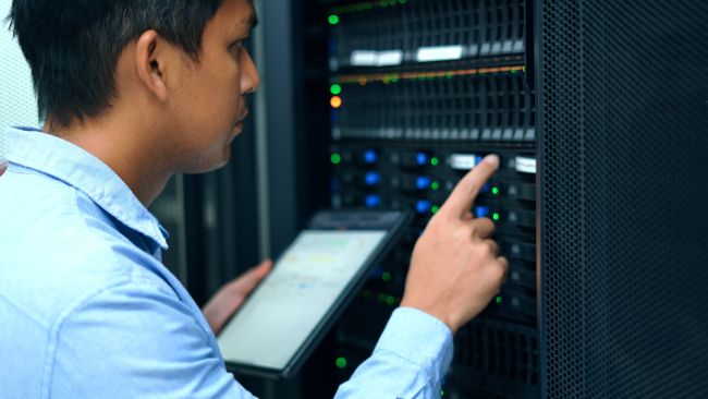 An illustrative image portrays an engineer meticulously checking a large server unit, aligning with the videoconference theme "Precision Automation: Cross-Connectivity and Integration into Workcells." The engineer, equipped with a laptop, is engaged in configuring and monitoring the server, which is pivotal in managing the data flow across a smart manufacturing production line. The server's lights and cables are visible, symbolizing the complex network and the seamless exchange of information between different parts of the production line, including robots, machine tools, and workcells. This visual conveys the critical role of technology and skilled professionals in ensuring efficient, interconnected operations within modern automated manufacturing environments.